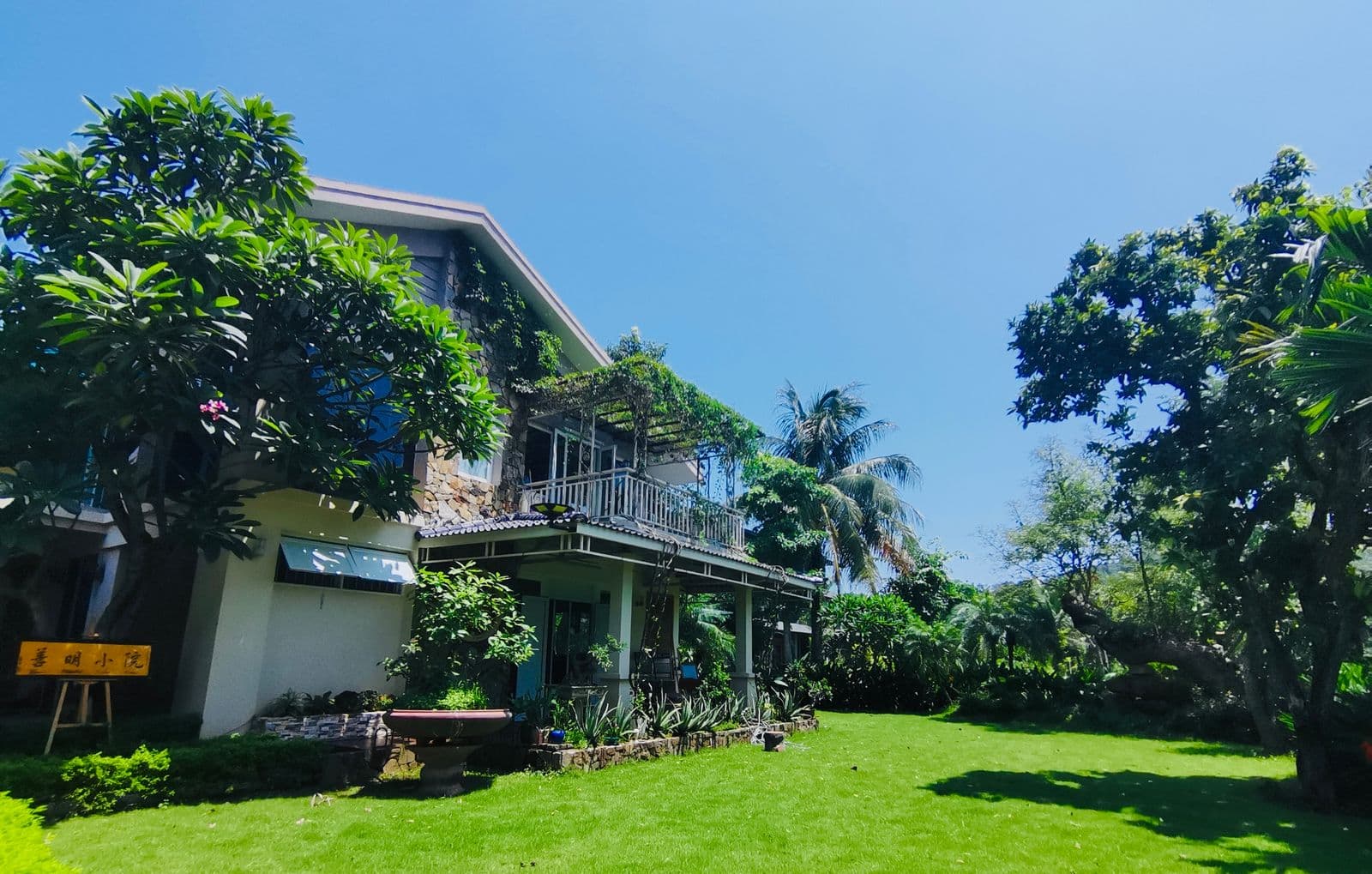 Two-story white building with upper balcony surrounded by lush tropical garden, palm trees, and green lawn under clear blue sky